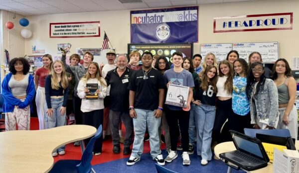 Steve Waddell poses with students in a classroom, holding a copy of his book "Valuepreneurs," surrounded by engaged and smiling students.